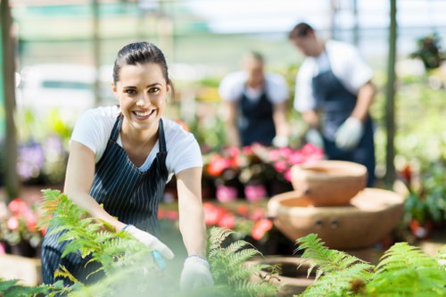 A smiling woman dressed in a white t-shirt and striped apron working in a lush, well-maintained greenhouse garden with abundant greenery and vibrant flowering plants. She is leaning forward slightly, tending to a bed of ferns with bright green fronds, while wearing gardening gloves. In the background, two other gardeners, a man and a woman, are engaged in planting or arranging flowers amidst rows of potted plants and floral displays; they are slightly out of focus. The greenhouse structure features transparent glass panels allowing natural sunlight to illuminate the scene, with glimpses of blue sky visible outside. The environment appears clean, organized, and dedicated to professional garden and landscaping maintenance, aligning with services provided by Lawn Mowing Haringey in North London. The scene emphasizes healthy plant growth, vibrant colours, and an active, professional gardening environment suitable for outdoor care and garden landscaping projects.