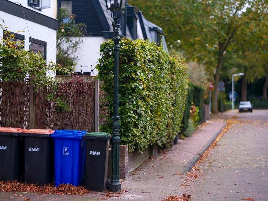A black wheeled garden waste bin filled with trimmed grass, dried ornamental grasses, and leafy plant matter is positioned on a grassy area in a garden. Surrounding the bin are lush green shrubs, a ye